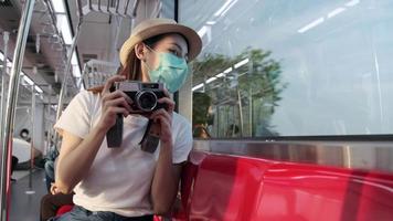 una hermosa turista asiática con mascarilla se sienta en un asiento rojo, viaja en tren, toma una foto instantánea, se transporta a la vista de los suburbios, disfruta del estilo de vida de los pasajeros en tren, feliz viaje de vacaciones. video