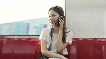 Beautiful Asian female tourist with camera sits in red seat, traveling by sky train, talking mobile phone when transporting in urban view, city passenger lifestyle by railway, happy journey vacation. video