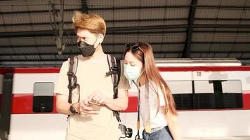 una pareja de turistas asiáticos con máscaras faciales busca información, encuentra lugares de viaje mediante un mapa de teléfono móvil en una estación de tren en tailandia, estilo de vida de viaje de pasajeros, transporte informal, vacaciones de viaje. video