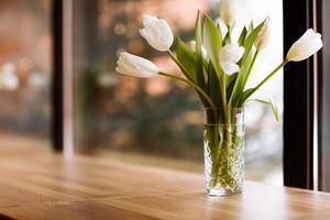 Vase with white flowers on big windows background with wooden table. Home coziness concept. Bouquet of tulips in glass vase.