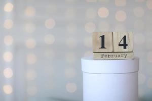 Valentine's day concept. Date 14 February on wooden cube calendar on a white gift box and white heart in hand. Bokeh golden and white background photo