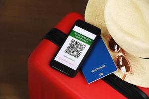 New normal concept. Close up view of a smartphone with an immune digital health passport, a red suitcase, protective mask, straw hat, passport and sunglasses. Traveling by plane during a pandemic. photo