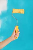 Cerca de la mano femenina que sostiene el rodillo de pintura amarillo sobre fondo azul-reparación, construcción y concepto de herramientas de construcción. herramienta conveniente y versátil para pintar paredes.