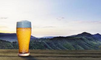 cerveza de barril o artesanal en vidrio transparente con espuma de cerveza en la parte superior puesta en un juego de mesas y sillas de madera. asientos de salón en la terraza de madera con fondo de montaña con luz de dios o luz solar.representación 3d foto