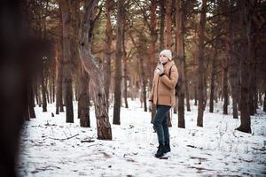 A young girl in winter clothes and a cup of hot drink walks in the winter forest. photo