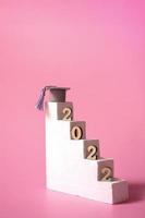 Simple concept of education on a pink background. Graduation 2022. Podium steps with graduation cap and wooden numbers closeup. Vertical Format photo