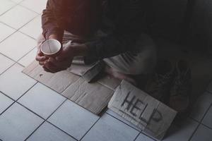 hombre adulto sin hogar sentado en la calle a la sombra del edificio y pidiendo ayuda y dinero con un cartel. concepto de personas sin hogar. foto