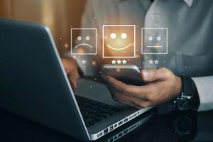 Business man holding phone wiht virtual screen on the happy Smiley face icon to give satisfaction in service. rating very impressed. Customer service and Satisfaction concept photo