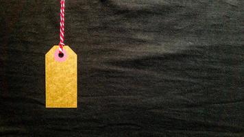 Brown cardboard tag hanging on a red rope on a black background from fabric. Copy space, place for text, flat lay. photo