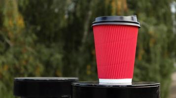 Coffee in a paper disposable eco glass of red color with a black plastic lid against the background of a green park. Selective focus. Close-up photo