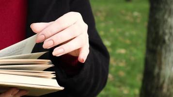 Girl reading a book in the park. Female hands flipping pages of paper book outdoors. The student is preparing for the exam. Literary leisure in nature. Close-up, copy space.