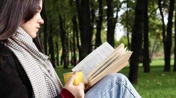 A woman sits under a tree and reads her favorite book while drinking coffee or tea from a yellow cup in a city park on green grass on a pleasant sunny day. Vacation, education and study concept. photo