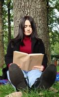 retrato de una hermosa joven que está sentada bajo un árbol y leyendo su libro favorito en un parque de la ciudad sobre hierba verde en un agradable día soleado. concepto de vacaciones, educación y estudio. foto