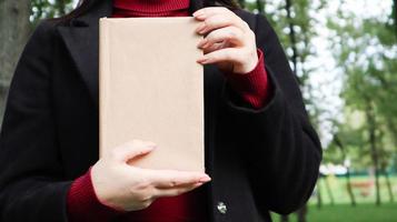 Close up view of woman in black woolen coat and burgundy sweater holding a book with empty cover in the park. Free space for your book reading concept background layout. photo