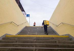 Ukraine, Kiev - September 26, 2019. A walking Glovo courier service provider with a yellow backpack rises the steps of the transition, hurries to deliver the order on time to the customer photo