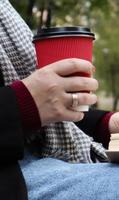 Young woman in jeans, coat and scarf, on a park bench. A woman is reading a book and drinking coffee or other hot drink outdoors alone. Close-up. The concept of honor, study, leisure and recreation. photo