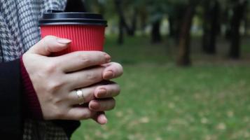 A young girl in a coat holds morning coffee with her while walking in the park. Hand holding paper cup of coffee in green park. Takeaway cappuccino. Close-up, copy space. photo