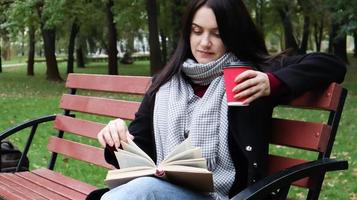 Young woman in jeans, coat and scarf, on a park bench. A woman is reading a book and drinking coffee or other hot drink outdoors alone. photo