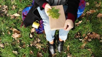 A woman holds a closed book lying on her lap with a fallen oak leaf in a park on a sunny warm autumn day on a green meadow. The concept of relaxation, reading and relaxation alone. Top view, flat lay. photo