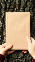 A closed book in a cover made of craft paper in female hands on the background of a tree bark in the park. Copy space. The concept of reading, recreation and leisure, study and education. photo