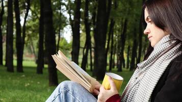 A woman sits under a tree and reads her favorite book while drinking coffee or tea from a yellow cup in a city park on green grass on a pleasant sunny day. Vacation, education and study concept. photo