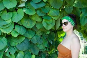 mujer joven sobre un fondo de un parque verde de verano, hojas verdes. una chica de pelo negro, una venda verde en la cabeza, con una camiseta marrón y gafas de sol negras foto