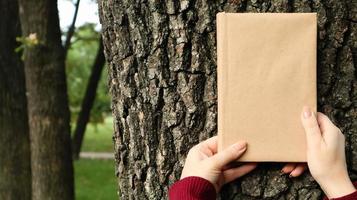 un libro cerrado en una cubierta hecha de papel artesanal en manos femeninas sobre el fondo de una corteza de árbol en el parque. copie el espacio el concepto de lectura, recreación y ocio, estudio y educación. foto