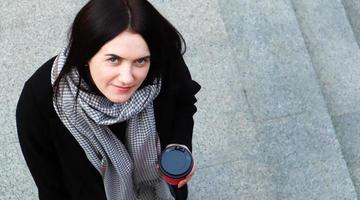 una joven mujer elegante con abrigo y bufanda bebe café caliente por la mañana en un vaso de papel ecológico rojo al aire libre en un parque de otoño. retrato de una mujer joven sosteniendo una taza de café para llevar, vista superior. foto