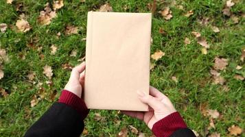 A closed book in a cover made of craft paper in female hands with green grass and fallen yellow leaves in the background. Flat lay, top view. Template, layout. Copy space. photo