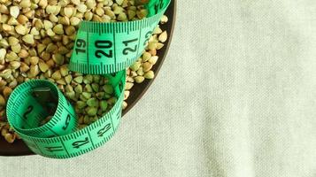 Green measuring tape on a background of raw buckwheat in a dish, concept of diet and healthy nutrition. Vegan organic food concept. Quarantine weight loss. Copy space for text. Top view, flat lay photo