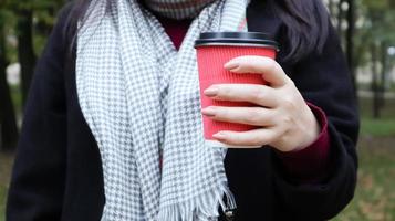 A young girl in a coat holds morning coffee with her while walking in the park. Hand holding paper cup of coffee in green park. Takeaway cappuccino. Close-up. photo