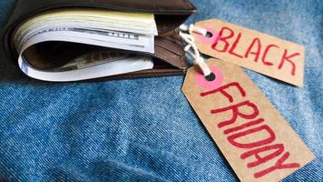 Brown wallet with money, dollars on a blue background, jeans texture, cardboard tag with red text Black Friday. Black Friday - International day of purchases, promotions, discounts, sales. photo