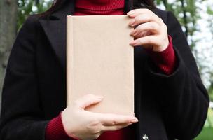 Close up view of woman in black woolen coat and burgundy sweater holding a book with empty cover in the park. Free space for your book reading concept background layout. photo