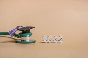 Graduation cap with stethoscope and numbers 2022 on beige background, closeup with copy space. Medical education concept. photo