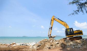 excavadora piedra excavadora trabajando en el sitio de construcción - retroexcavadora en la playa mar océano y fondo de cielo azul foto