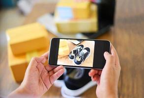 mujer joven tomando zapatillas fotográficas con un teléfono inteligente con cámara para publicar para vender en línea en el sitio web del mercado de Internet vendiendo en línea ecommerce envío compras en línea entrega y pedido foto