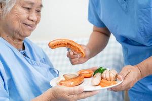 paciente asiática mayor o anciana anciana desayunando salchichas y vegetales alimentos saludables con esperanza y feliz mientras está sentada y hambrienta en la cama en el hospital. foto