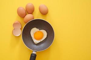 huevo frito en forma de corazón en la sartén con cáscara de huevo y huevos de gallina sobre fondo amarillo. vista superior. concepto de comida saludable. copiar espacio para texto foto