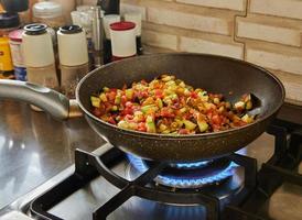 Dish with eggplant, zucchini and bell pepper is fried in pan on gas stove photo