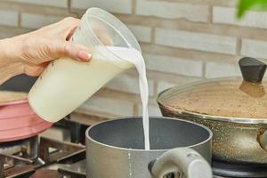 Woman pours milk from jug into pan for making cream pie in her home kitchen photo