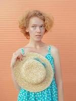 young cheerful curly redhead woman in blue sundress holding straw hat in her hand on beige background. Fun, summer, fashion, youth concept photo