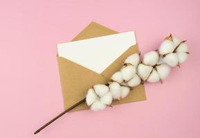 Blank card and vintage mail envelope on pink background with cotton flowers top view, space for text photo