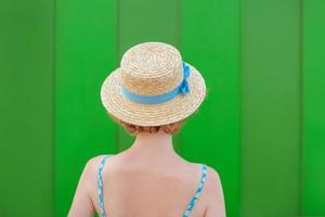 back of young curly redhead woman in straw hat and blue sundress standing on green wall background. Fun, summer, fashion, youth concept. Copy space