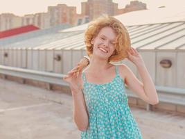 young happy cheerful curly redhead woman in straw hat, blue sundress eating ice cream on skyscraper roof. Fun, lifestyle, urban, modern, roof, city, summer, fashion, youth concept