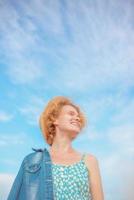 young curly redhead woman in straw hat, blue sundress and jeans jacket standing on blue sky background. Fun, summer, fashion, shooting, travel, youth concept. Copy spase