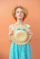 young cheerful curly redhead woman in blue sundress holding straw hat in her hand on beige background. Fun, summer, fashion, youth concept