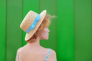 back of young curly redhead woman in straw hat and blue sundress standing on green wall background. Fun, summer, fashion, youth concept. Copy space photo