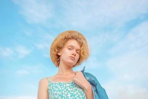 young curly redhead woman in straw hat, blue sundress and jeans jacket standing on blue sky background. Fun, summer, fashion, shooting, travel, youth concept. Copy spase