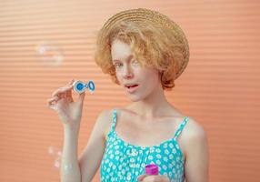 young cheerful curly redhead woman in blue sundress and straw hat with bubbles on beige background. Fun, summer, fashion, youth concept photo