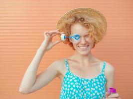 young cheerful curly redhead woman in blue sundress and straw hat with bubbles on beige background. Fun, summer, fashion, youth concept photo